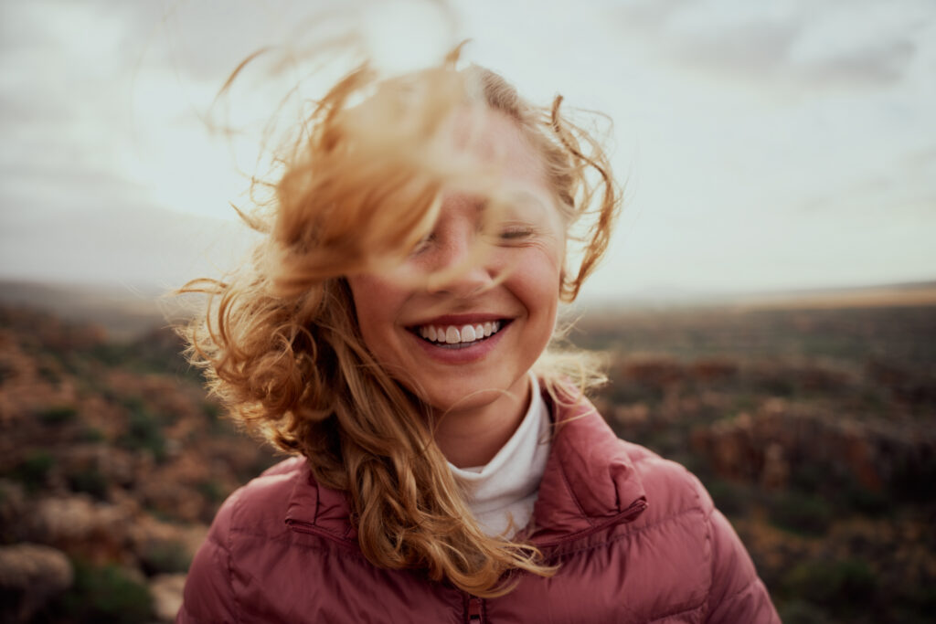 Eine junge Frau steht bei windigem Wetter lachend in der Natur