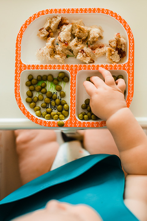 Das Foto zeigt ein Kleinkind in seinem Babystuhl mit einem Teller Essen vor sich.