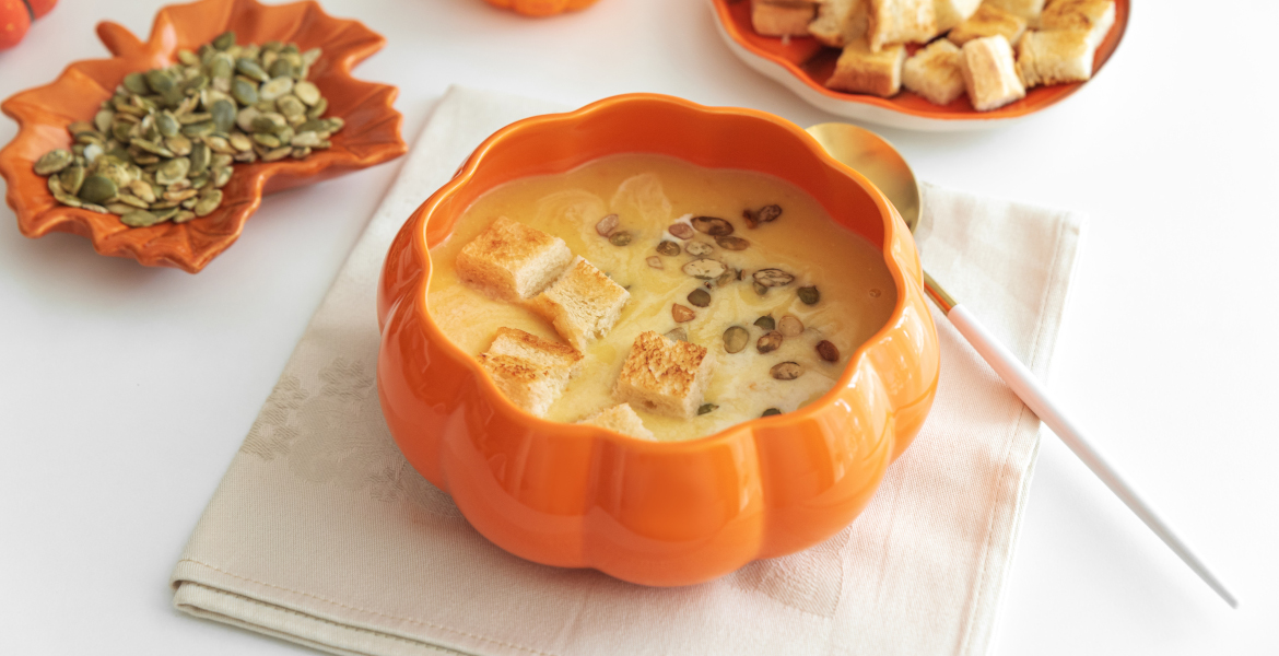 Cremige Kürbissuppe mit Croutons und Samen, serviert in einer kürbisförmigen Schüssel auf einem weißen Holztisch.